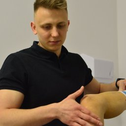 Fizjoterapeuta Bartosz Drobina fizjoterapia i rehabilitacja - Młody mężczyzna w czarnej koszulce polo bada kolano pacjenta w białej skarpetce z niebieskimi paskami, w jasnym gabinecie.