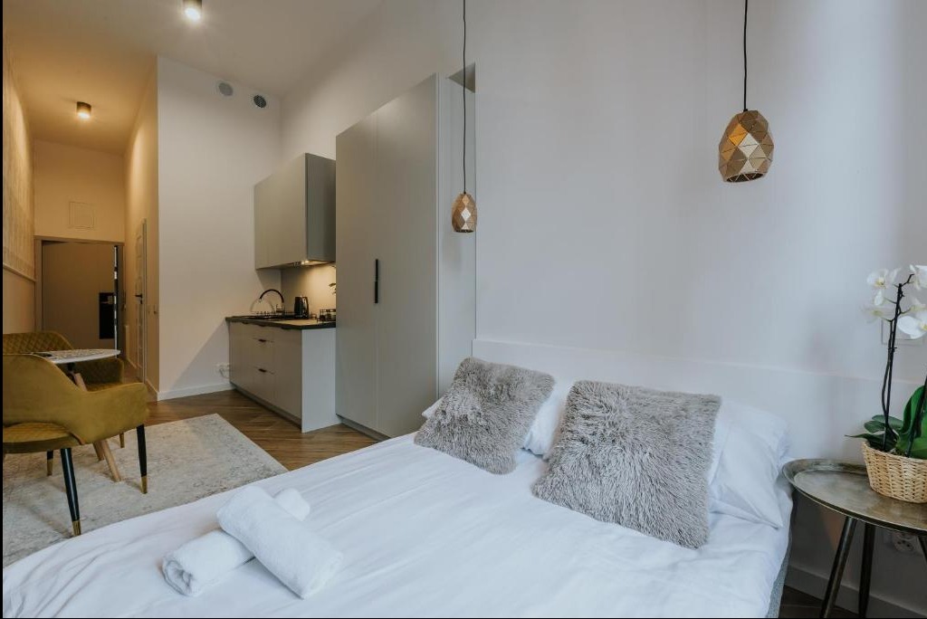 Nowoczesne studio z aneksem kuchennym i łóżkiem, szare poduszki dekoracyjne, lampy wiszące o geometrycznym kształcie, białe ściany, orchidea w koszyku na stoliku nocnym.