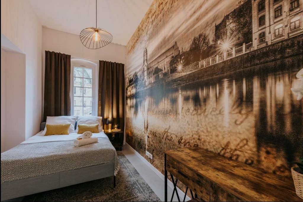 Pokój z łóżkiem, lampą wiszącą i fototapetą przedstawiającą panoramę miasta z rzeką, utrzymaną w sepii.