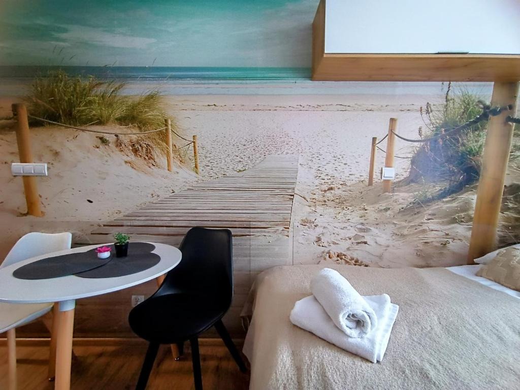 Pokój z fototapetą przedstawiającą plażę z drewnianą ścieżką, stolik z dwoma krzesłami i małą roślinką, oraz łóżko z ręcznikami