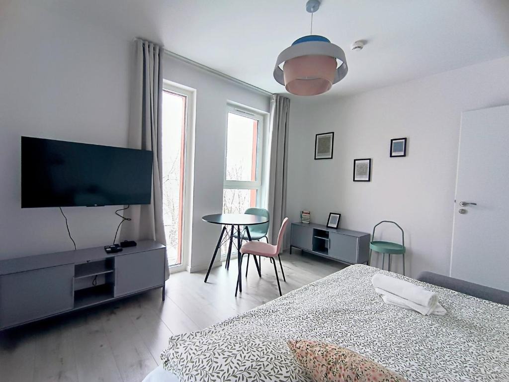 Jasne, minimalistyczne wnętrze pokoju z łóżkiem w kwiatowy wzór, szarym stolikiem RTV pod telewizorem, okrągłym stolikiem z krzesłami i wiszącą lampą z elementami różu i błękitu.