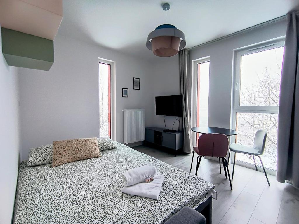 Jasne wnętrze pokoju z łóżkiem w zielono-białą pościel, stolikiem z krzesłami przy oknie i telewizorem na ścianie.