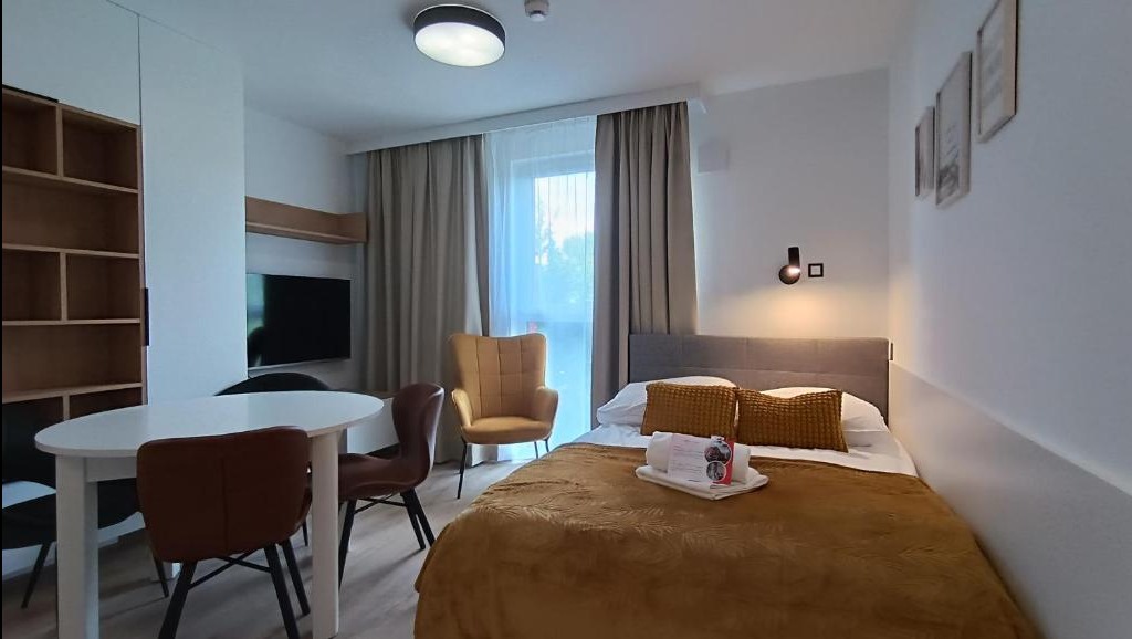 Pokój hotelowy z łóżkiem, stolikiem, krzesłami i telewizorem, utrzymany w stonowanej kolorystyce z akcentami w odcieniach brązu i beżu.