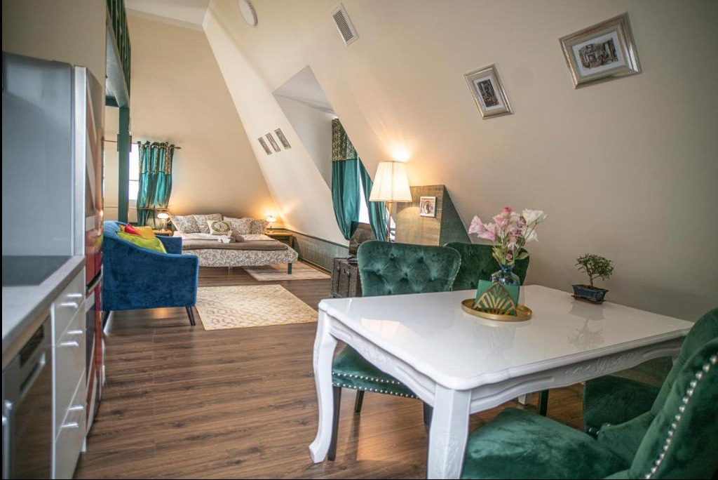 Jasne poddasze ze skośnymi ścianami, urządzone w stylu eklektycznym: biały stół z zielonymi krzesłami, niebieski fotel, łóżko z wzorzystą pościelą i turkusowymi zasłonami, drewniana podłoga.