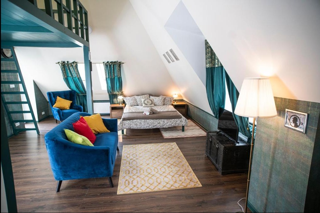 Jasne poddasze z antresolą, niebieską sofą z kolorowymi poduszkami, łóżkiem małżeńskim, dywanem w geometryczne wzory i lampą podłogową