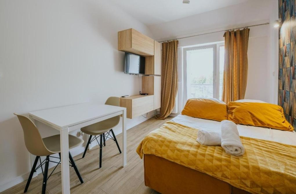 Jasne wnętrze pokoju z drewnianą podłogą, białym stołem z krzesłami, łóżkiem z żółtą pościelą i zwiniętymi ręcznikami, oraz wiszącą szafką z telewizorem na ścianie.