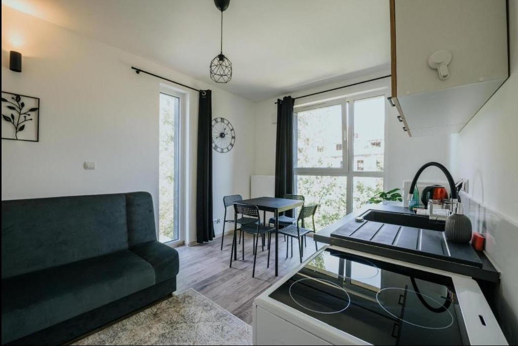 Nowoczesne wnętrze kawalerki: ciemnozielona sofa, czarny stół z krzesłami, aneks kuchenny z płytą indukcyjną, okna z czarnymi zasłonami, dekoracyjny zegar na ścianie.