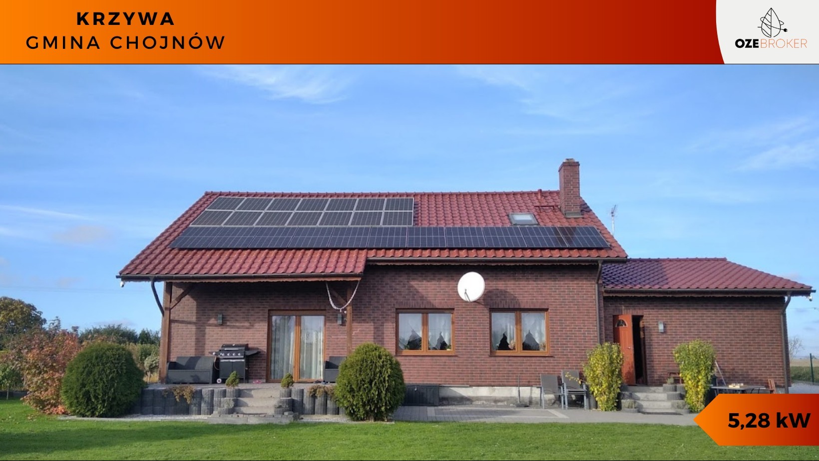 Ceglany dom jednorodzinny z czerwonym dachem, na którym zamontowano panele słoneczne; widoczna antena satelitarna i zadbany trawnik.
