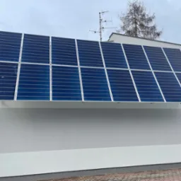 Instalacja paneli słonecznych na dachu budynku z szarą elewacją, widoczna antena telewizyjna i drzewa w tle.