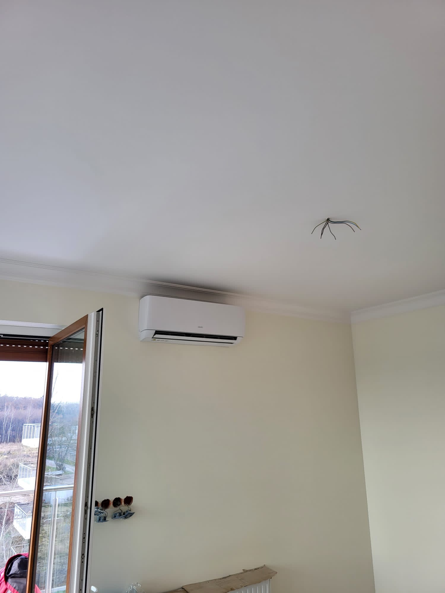 Wewnętrzna jednostka klimatyzacji zamontowana na ścianie nad grzejnikiem, widoczne wystające przewody elektryczne na suficie oraz puszki elektryczne bez osprzętu przy oknie balkonowym.