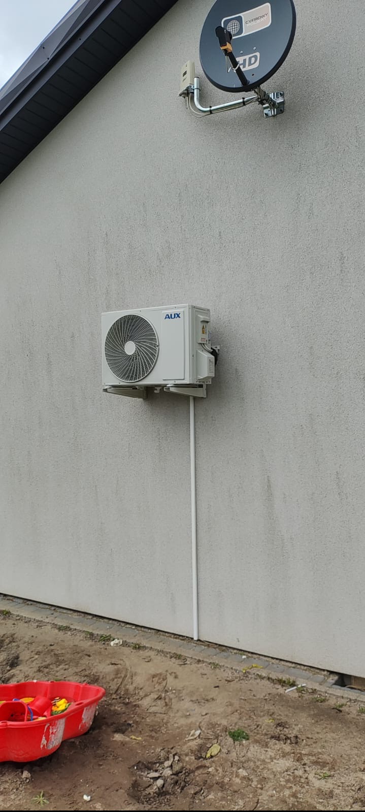 Zewnętrzna jednostka klimatyzacji marki AUX zamontowana na elewacji domu z widoczną rurą odprowadzającą i anteną satelitarną powyżej, u dołu widoczna piaskownica.