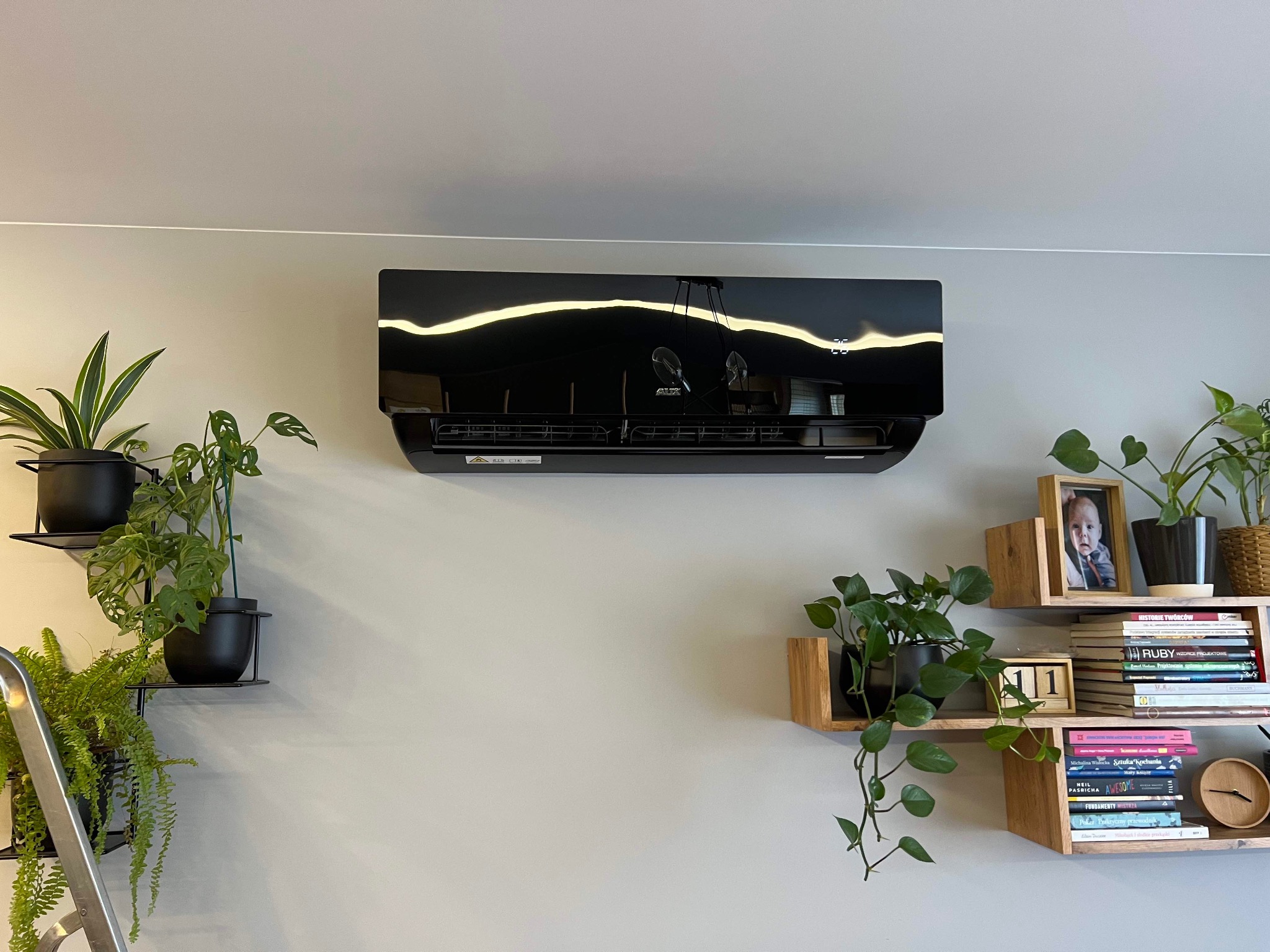 Czarny klimatyzator ścienny zamontowany w jasnym pomieszczeniu, otoczony roślinami doniczkowymi na półkach i drabina oparta o ścianę.