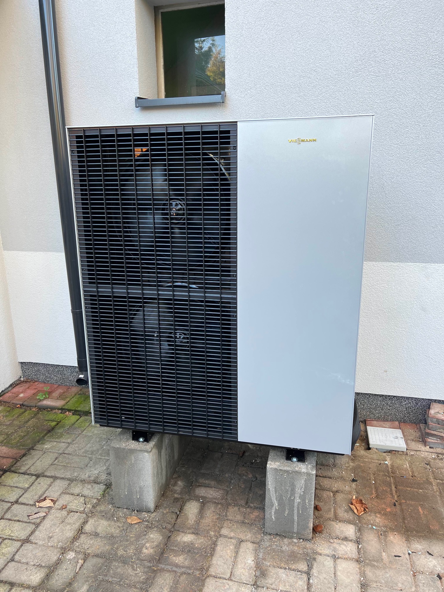 Zewnętrzna jednostka pompy ciepła Viessmann zamontowana na betonowych postumentach przy ścianie budynku z widoczną rynną spustową.