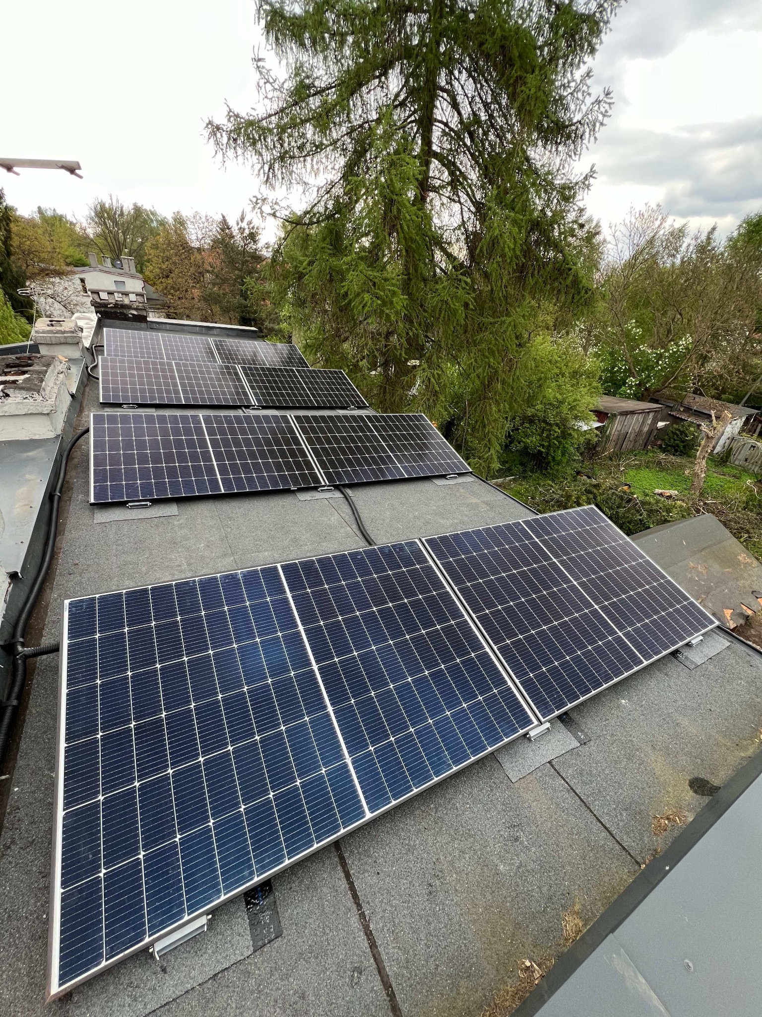 Panele fotowoltaiczne zamontowane na dachu pokrytym papą, widoczne okablowanie i otaczająca zieleń drzew.