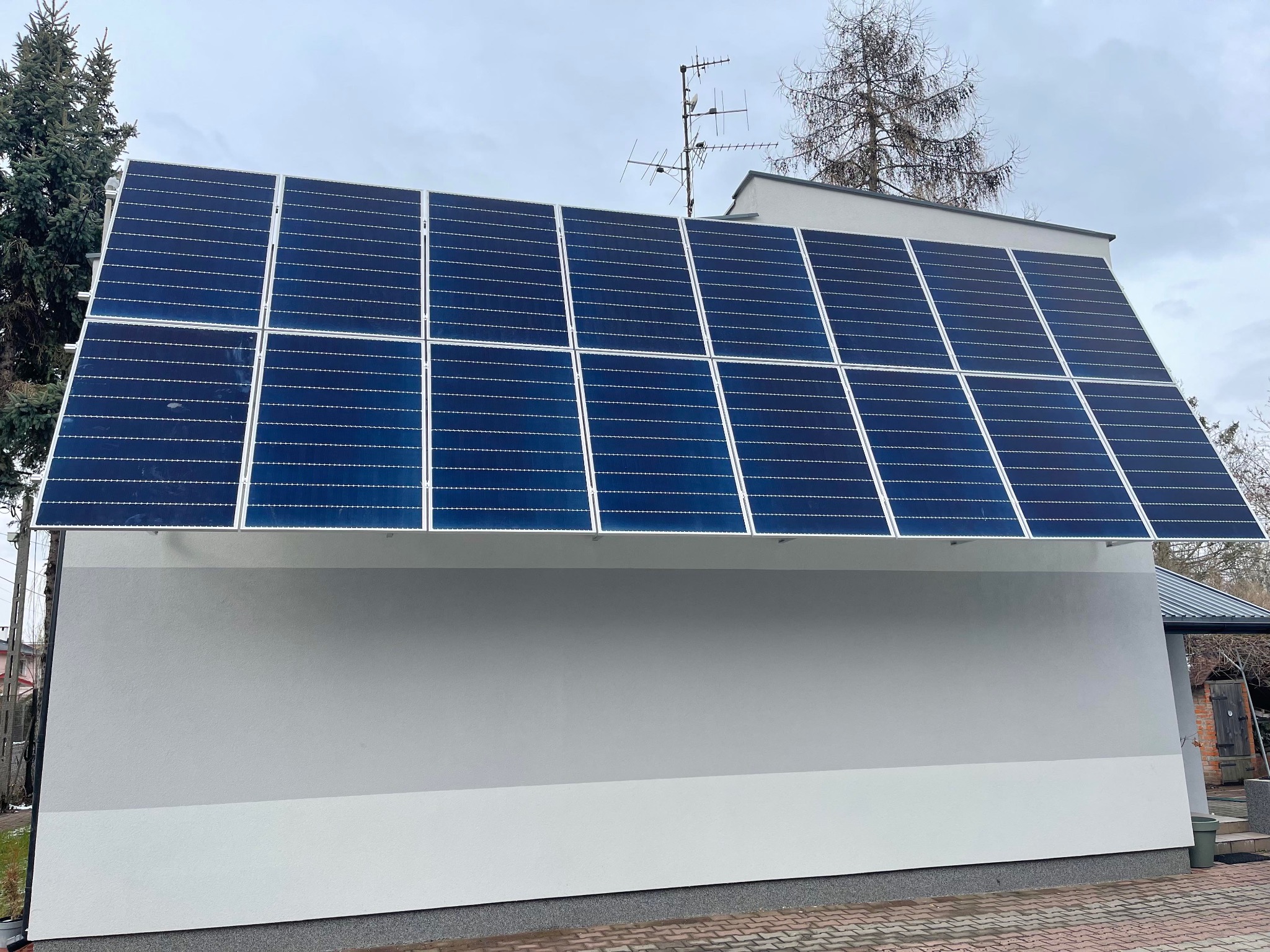 Instalacja paneli słonecznych na dachu budynku z szarą elewacją, widoczna antena telewizyjna i drzewa w tle.