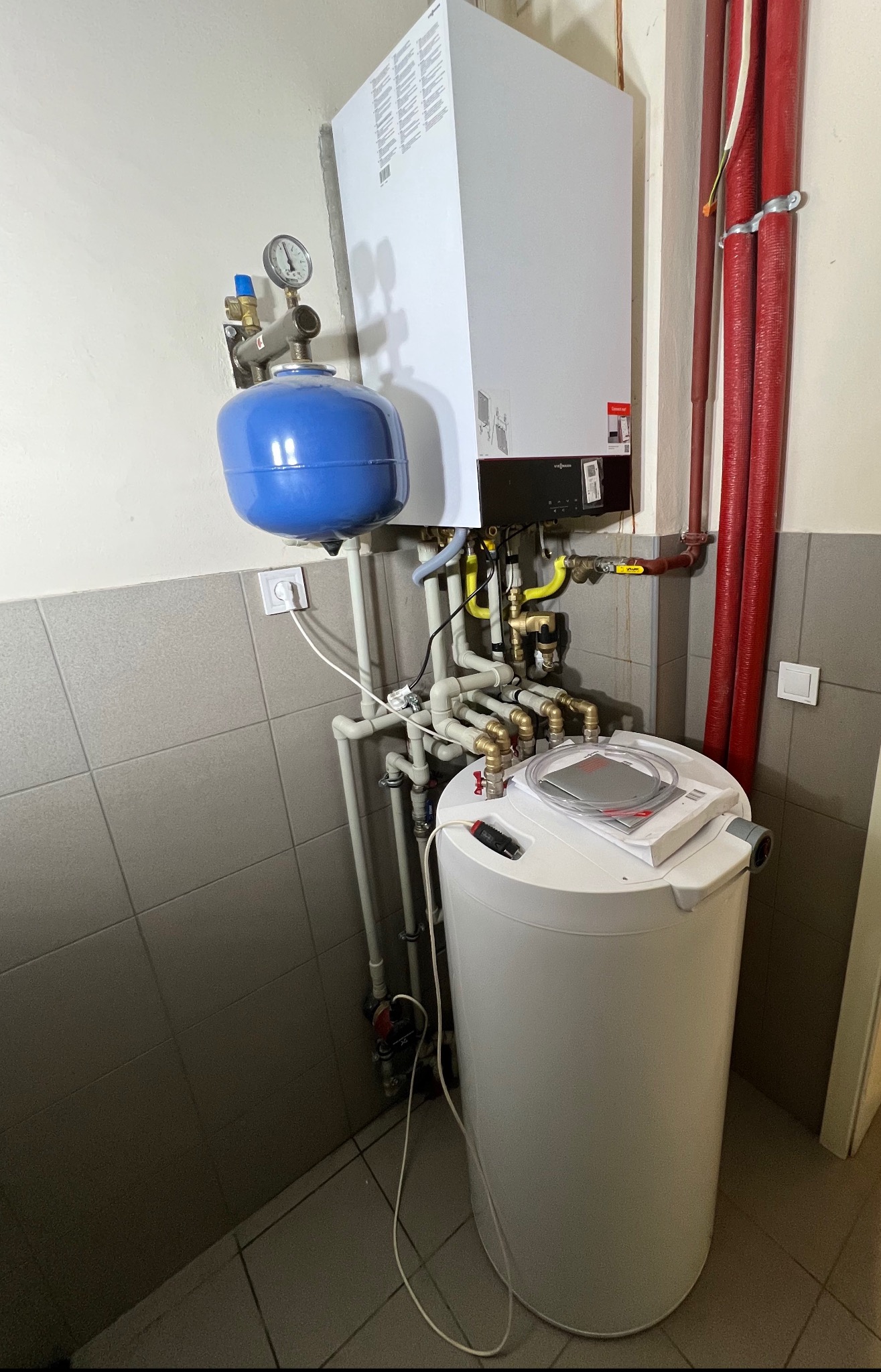Zestaw grzewczy z piecem gazowym Viessmann, zbiornikiem i niebieskim naczyniem przeponowym, widoczne rury, zawory i manometr, montaż w pomieszczeniu z szarymi płytkami.