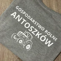 Szara koszulka z białym nadrukiem nazwy 'Gospodarstwo Rolne Antoszków' i grafiki traktora, leżąca na drewnianym podłożu.