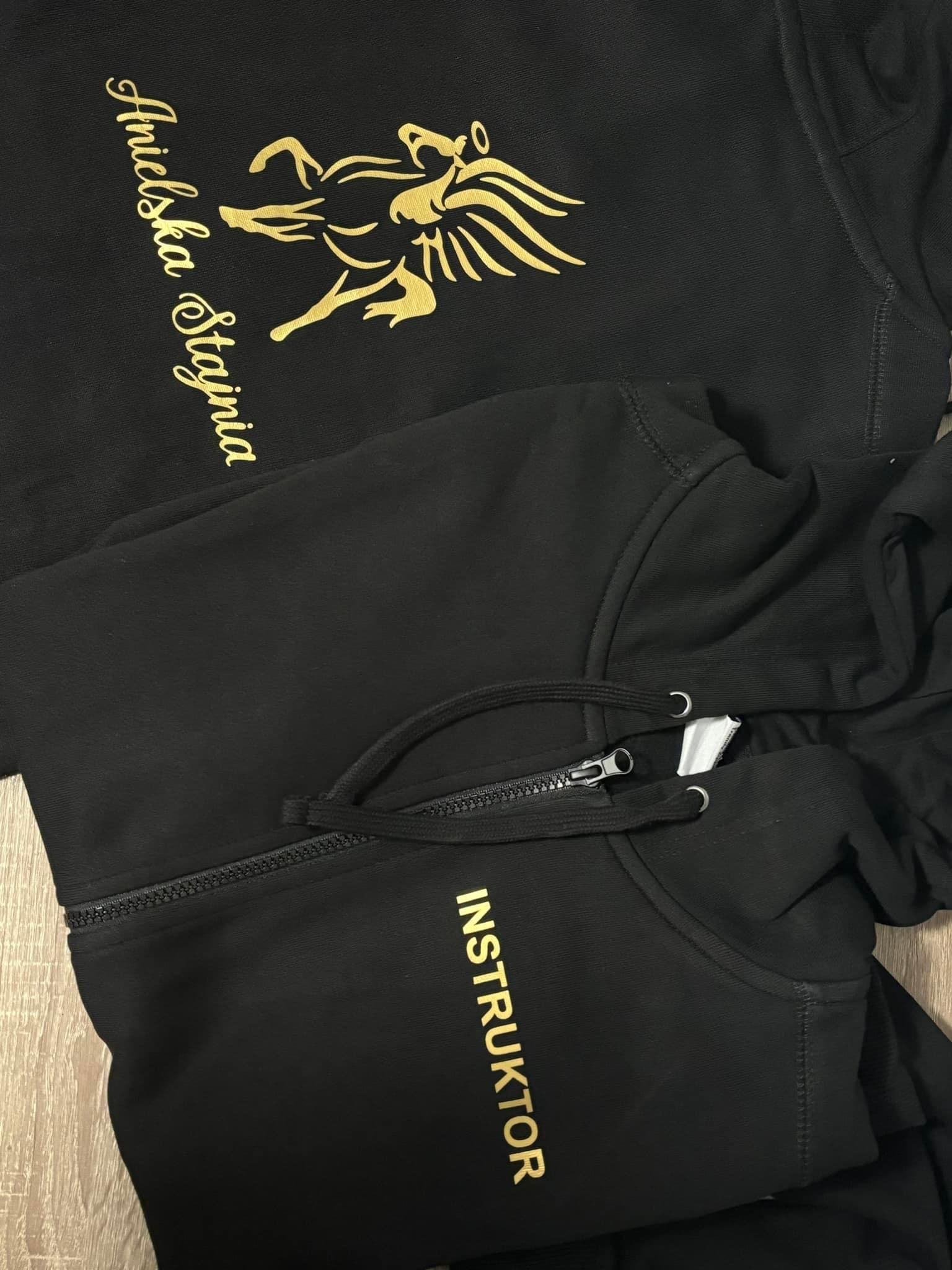 Czarne bluzy z kapturem: jedna z nadrukiem 'Anielska Stajnia' i złotym logo, druga z napisem 'INSTRUKTOR' w złotym kolorze, widoczny zamek błyskawiczny.