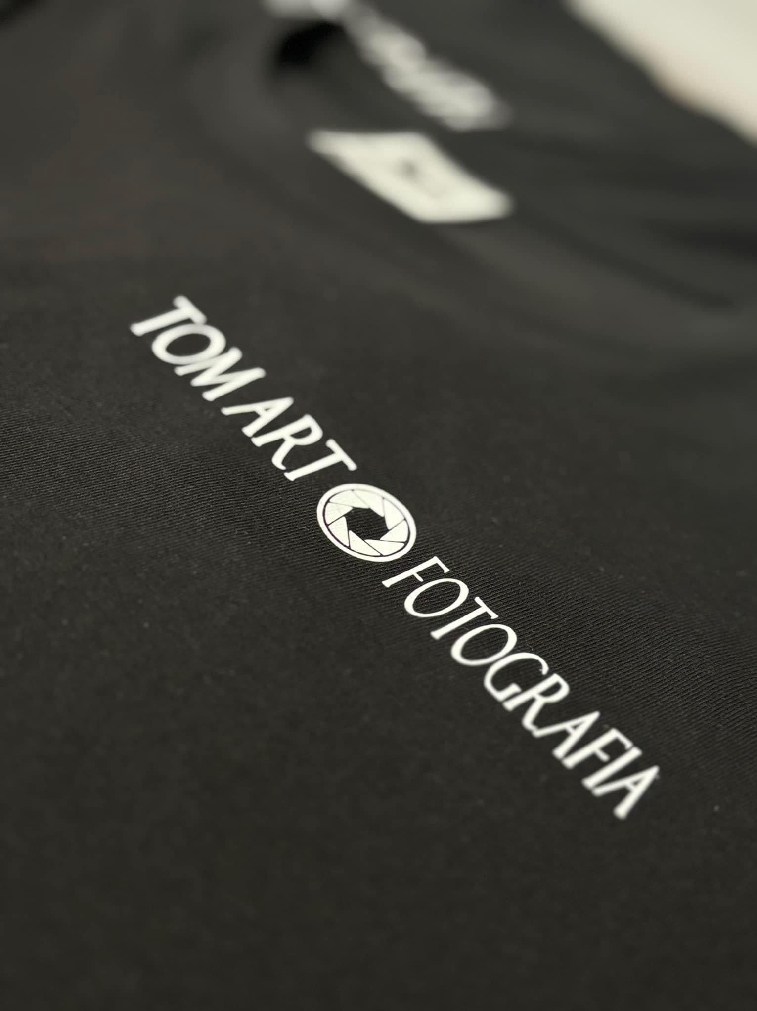 Czarna koszulka z białym nadrukiem 'TOM ART FOTOGRAFIA' z symbolem przysłony aparatu, widoczna faktura materiału pod kątem.