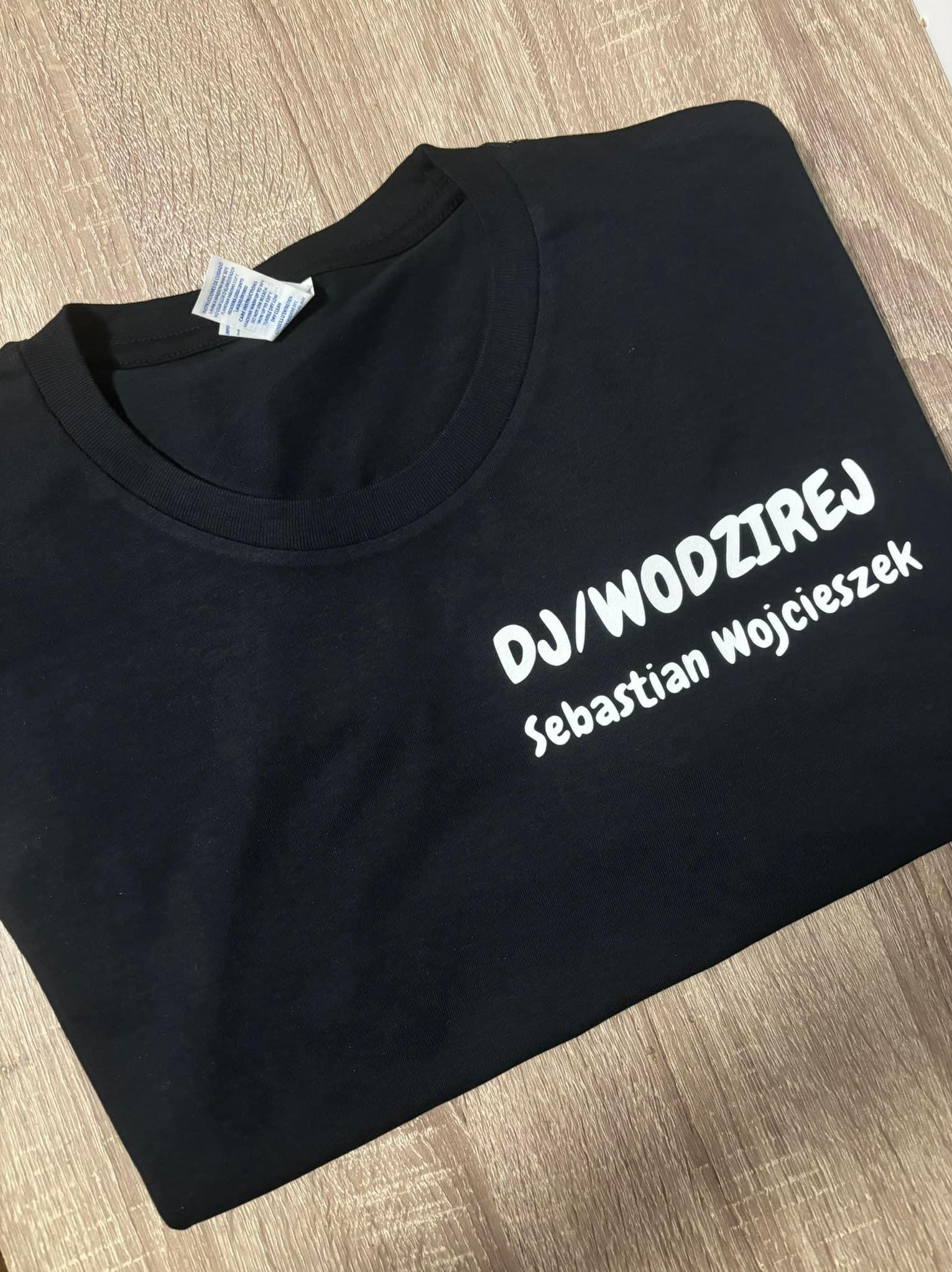 Czarna koszulka z białym napisem 'DJ/WODZIREJ Sebastian Wojcieszek' leżąca na drewnianym podłożu, widoczna metka wszyta w kołnierzyk.