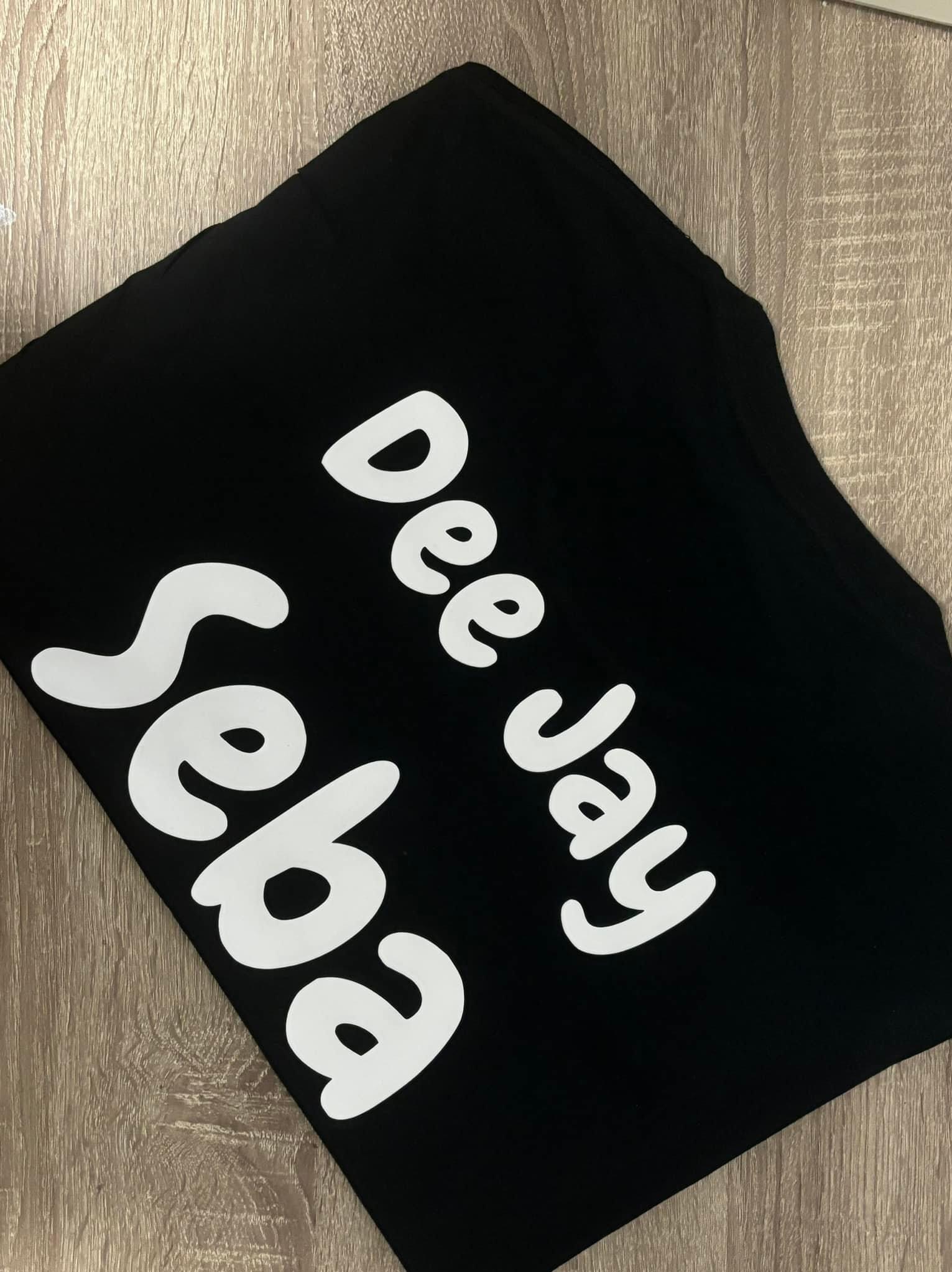 Czarna koszulka z białym nadrukiem 'Dee Jay Seba' leżąca ukośnie na jasnym, drewnianym tle.