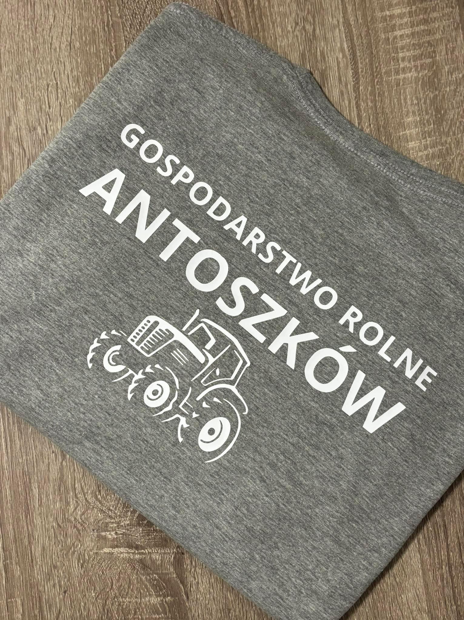 Szara koszulka z białym nadrukiem nazwy 'Gospodarstwo Rolne Antoszków' i grafiki traktora, leżąca na drewnianym podłożu.