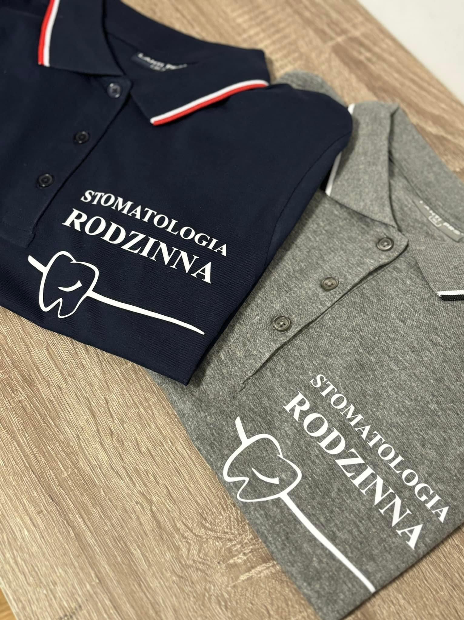 Dwa polo w kolorze granatowym i szarym z nadrukiem 'Stomatologia Rodzinna' i stylizowanym zębem, leżące na drewnianym blacie.