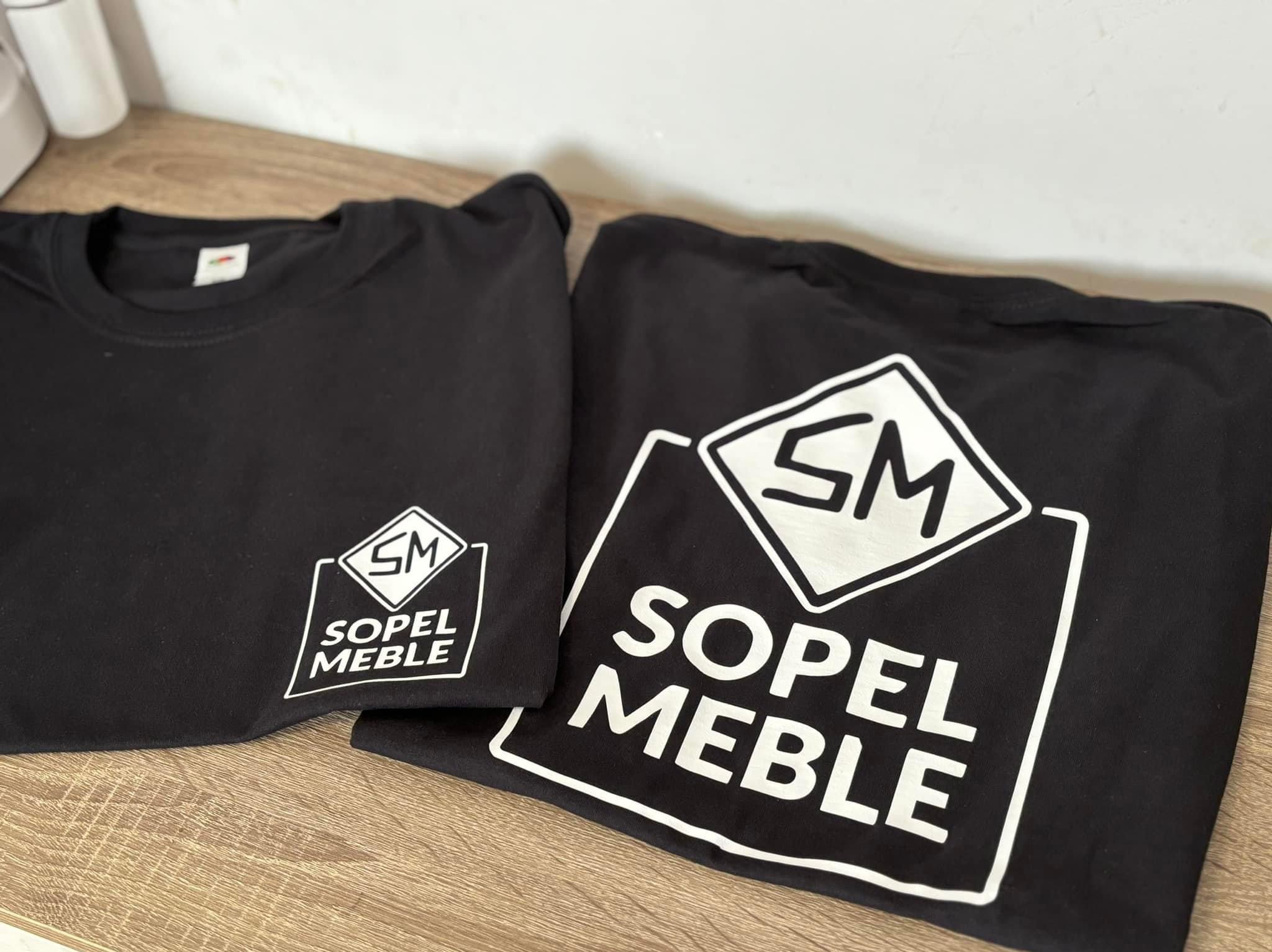 Dwie czarne koszulki z białym nadrukiem logo firmy Sopel Meble, na jednej z nich logo jest większe.