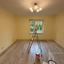 Pokój po malowaniu ścian na jasnożółty kolor. Na podłodze folia ochronna, drabina, pędzle i farby Dekoral. Widoczne okno z widokiem na zieleń.