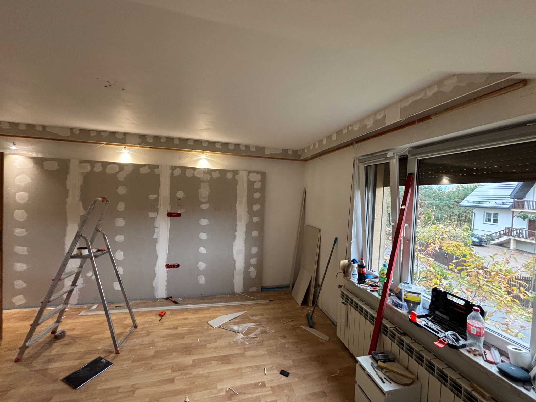 Pokój w trakcie remontu: ściany z płyt gipsowo-kartonowych, częściowo zaszpachlowane, drabina, narzędzia na parapecie. Widok na zewnątrz przez okno.