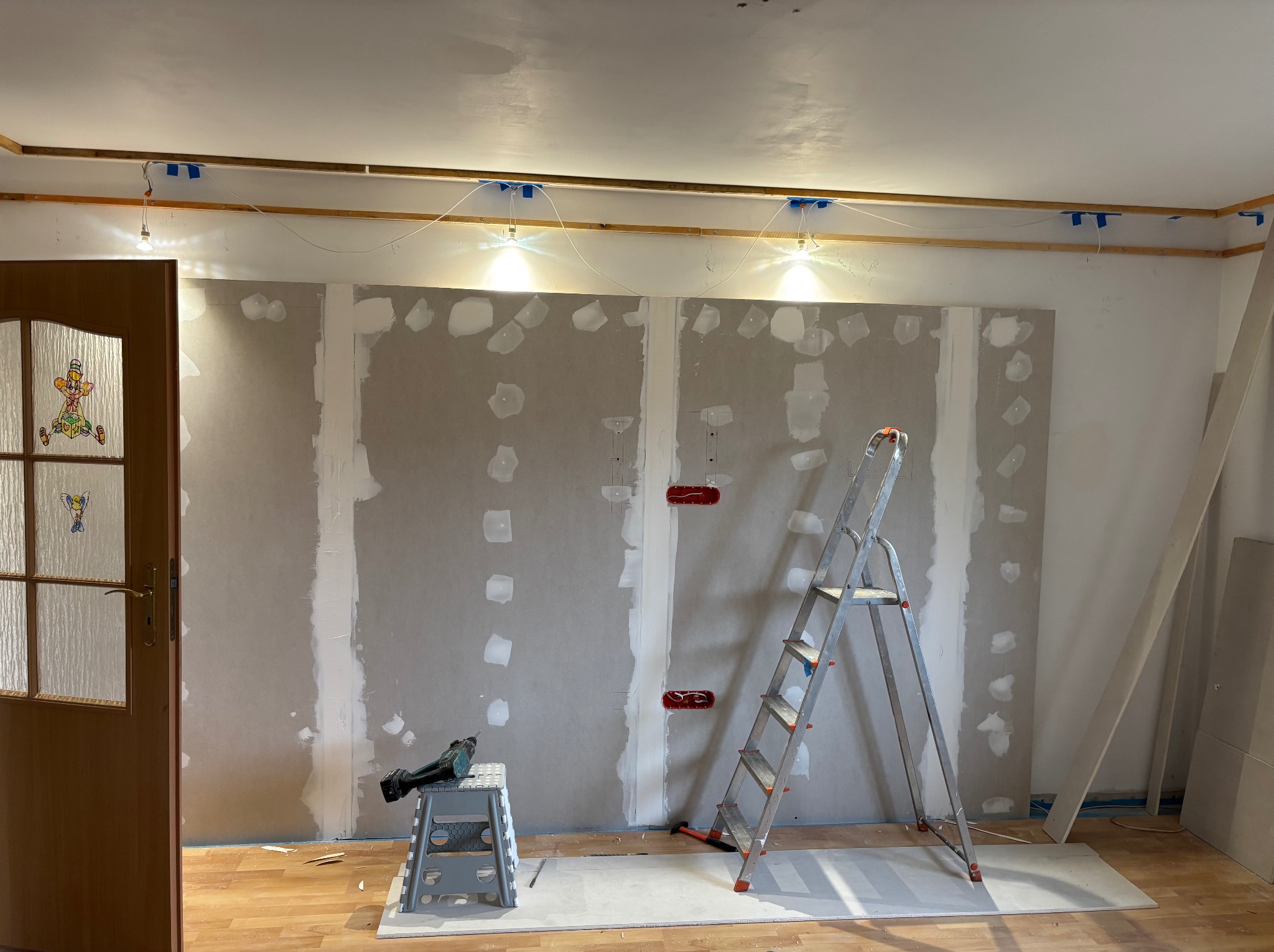 Montaż płyt gipsowo-kartonowych na ścianie z widocznymi puszkami elektrycznymi, drabina i narzędzia. Widoczne oświetlenie tymczasowe i drewniana konstrukcja pod sufitem.
