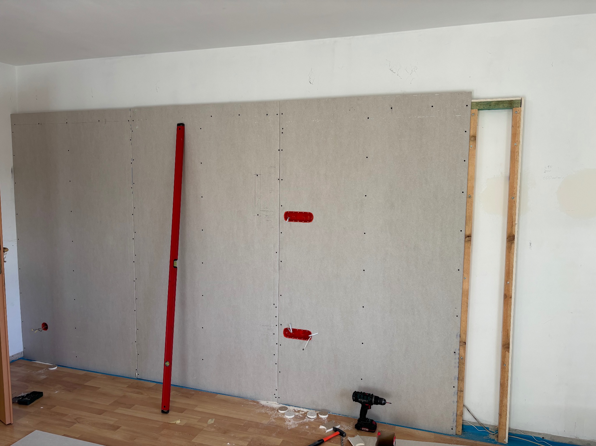Montaż płyt gipsowo-kartonowych na ścianie z widocznym poziomem, wiertarką i elementami instalacji elektrycznej. Prace remontowe w trakcie realizacji.