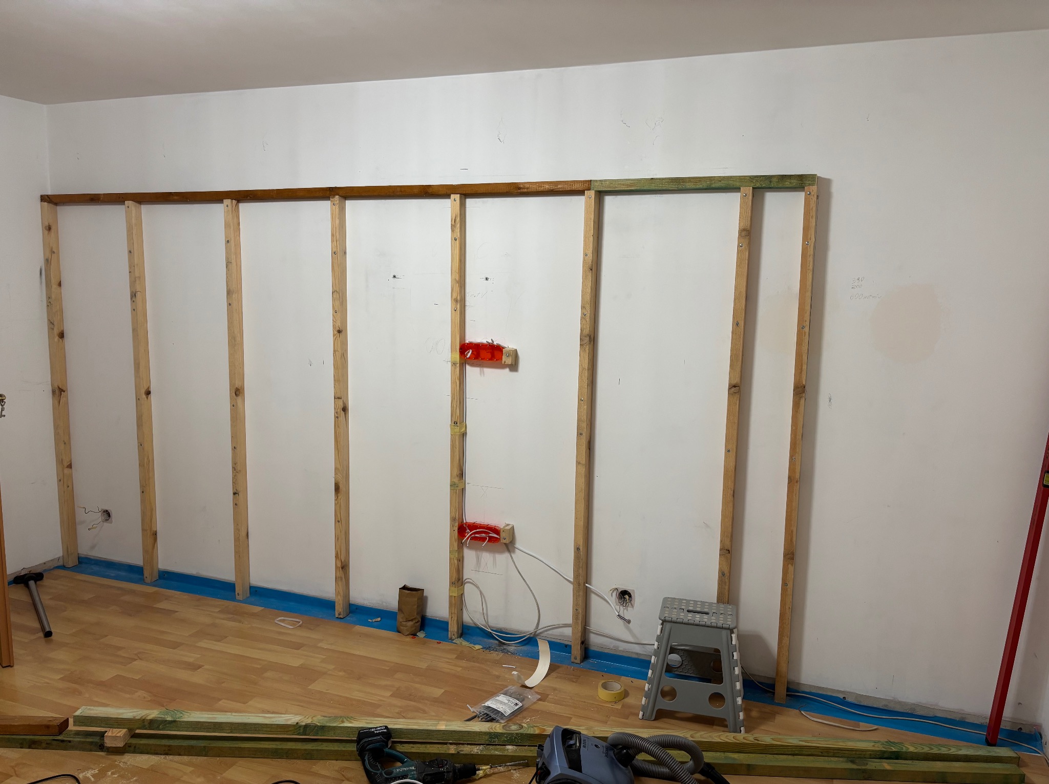 Konstrukcja szkieletowa ściany działowej z drewna, widoczne przewody elektryczne, narzędzia na podłodze. Etap budowy ściany w pomieszczeniu z panelami.