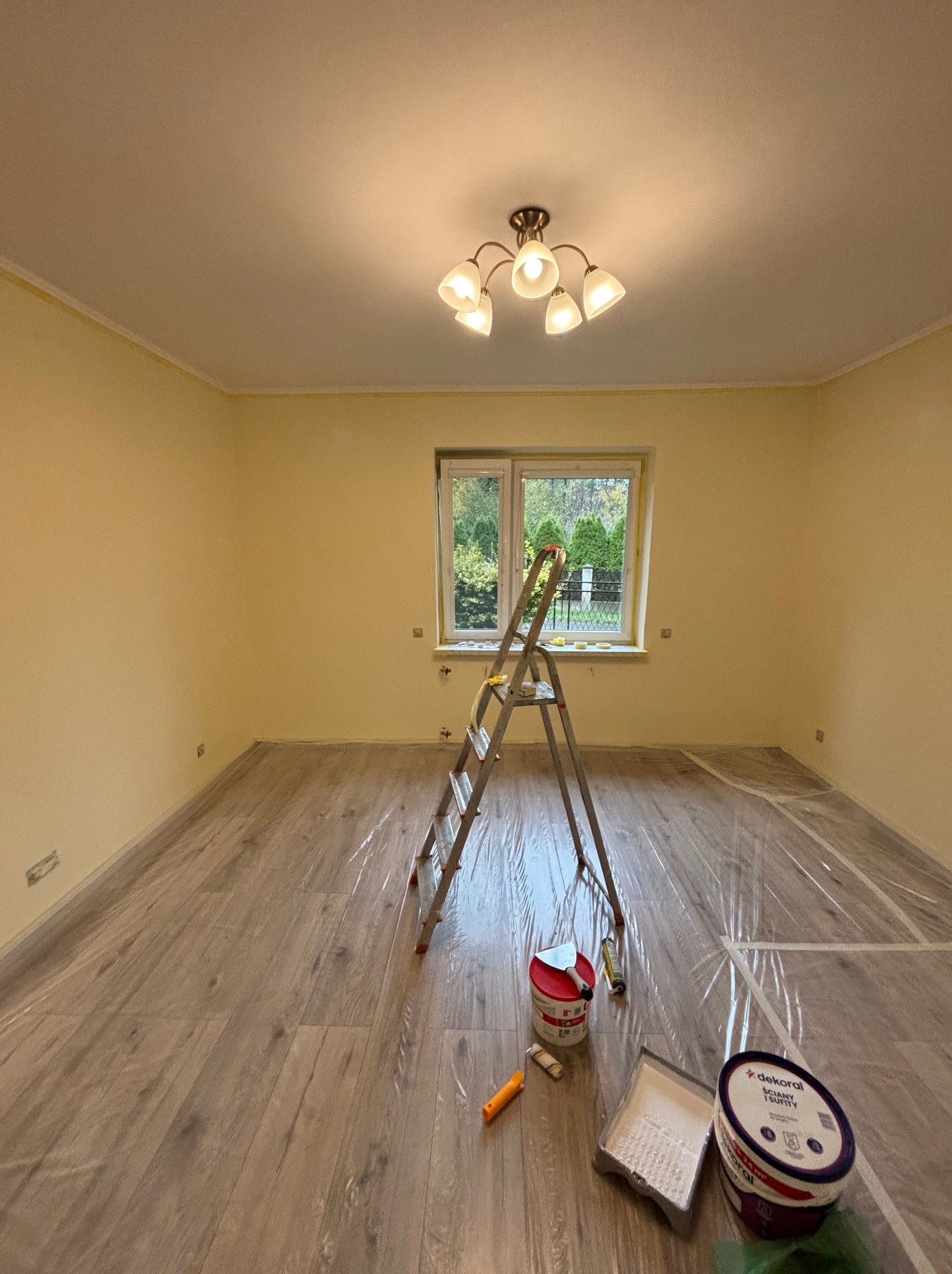 Pokój po malowaniu ścian na jasnożółty kolor. Na podłodze folia ochronna, drabina, pędzle i farby Dekoral. Widoczne okno z widokiem na zieleń.
