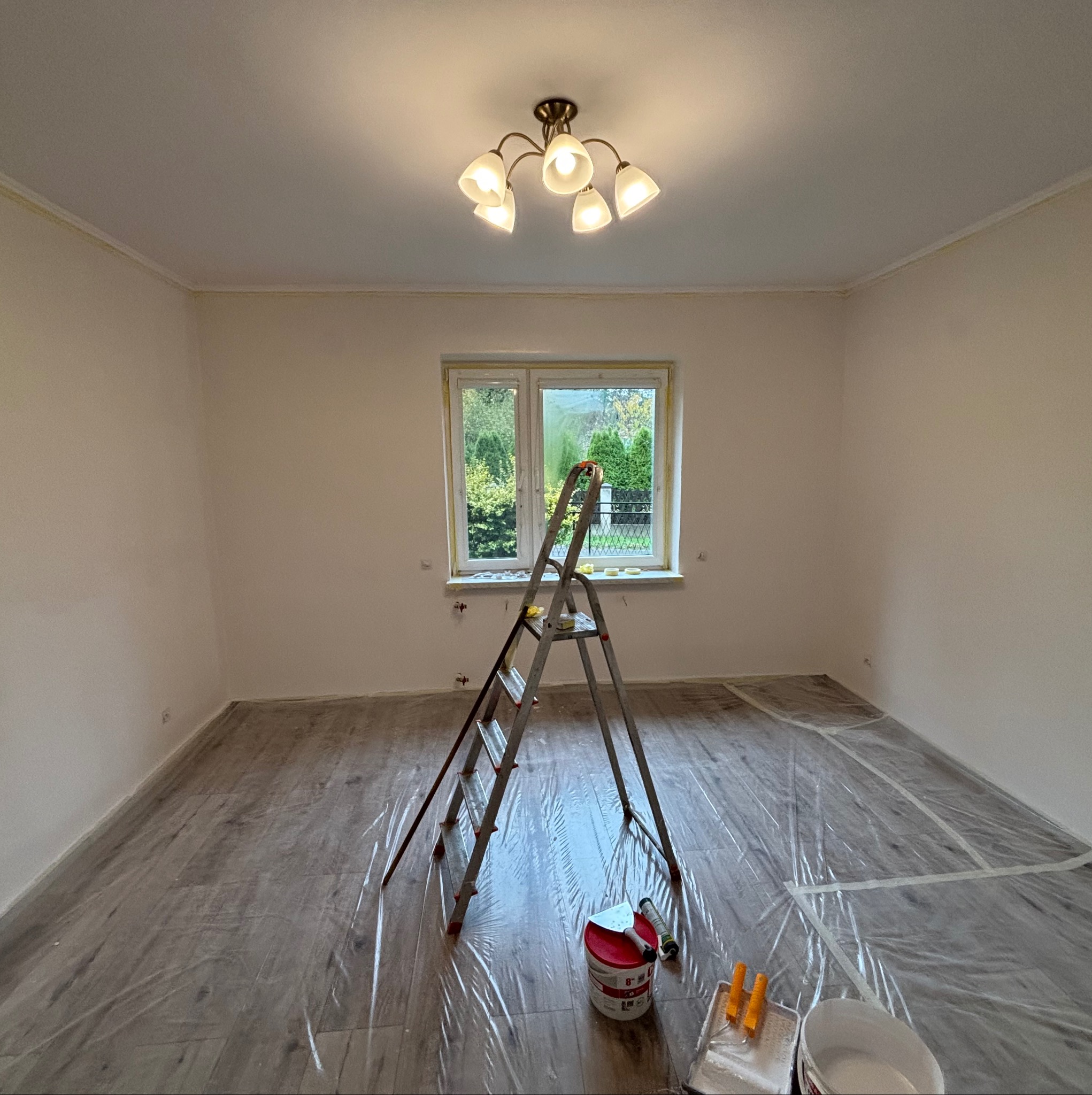 Pokój w trakcie malowania: drabina, farby, folia ochronna na podłodze. Widoczne przygotowania do malowania ścian w jasnym wnętrzu z oknem.