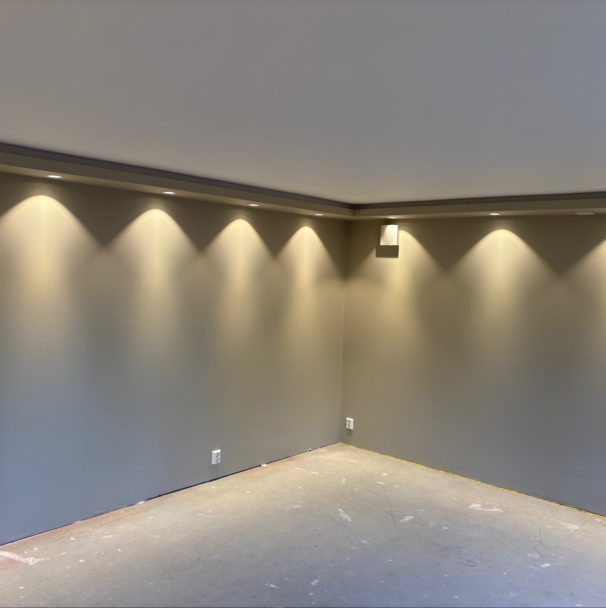 Szare ściany w pustym pokoju z oświetleniem punktowym LED na suficie. Widoczne gniazdka elektryczne i surowa podłoga przed wykończeniem.