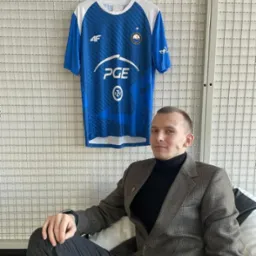 Mężczyzna w tweedowej marynarce siedzi na tle białej ściany z siatki, obok wisi niebieska koszulka sportowa z logo PGE i innymi nadrukami.