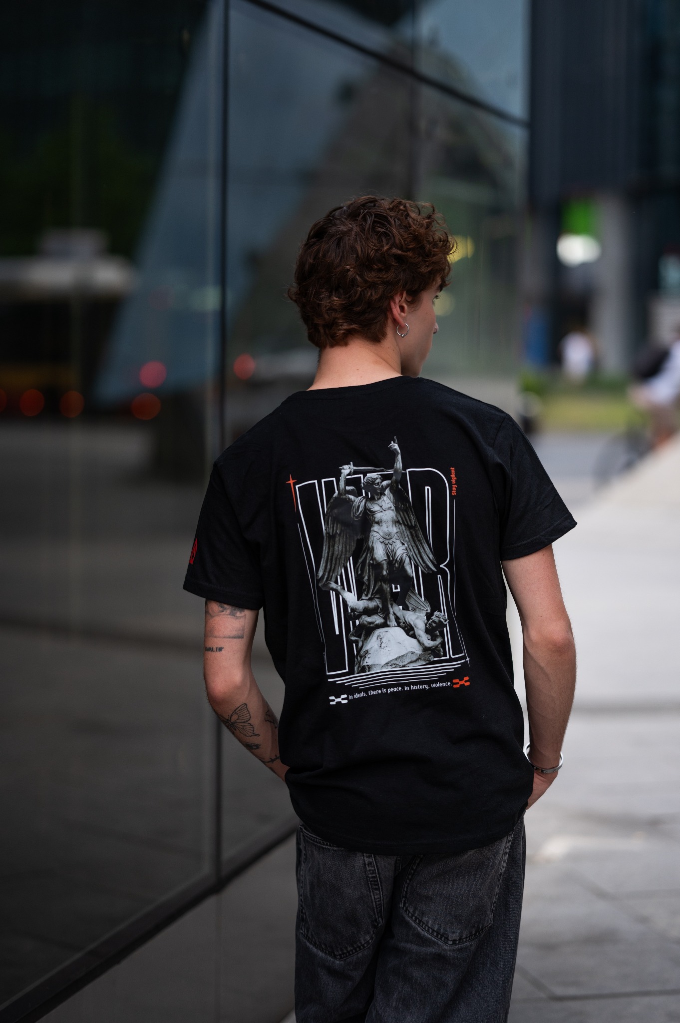 Mężczyzna w czarnym T-shircie z nadrukiem anioła i napisem, stojący tyłem na tle przeszklonego budynku.