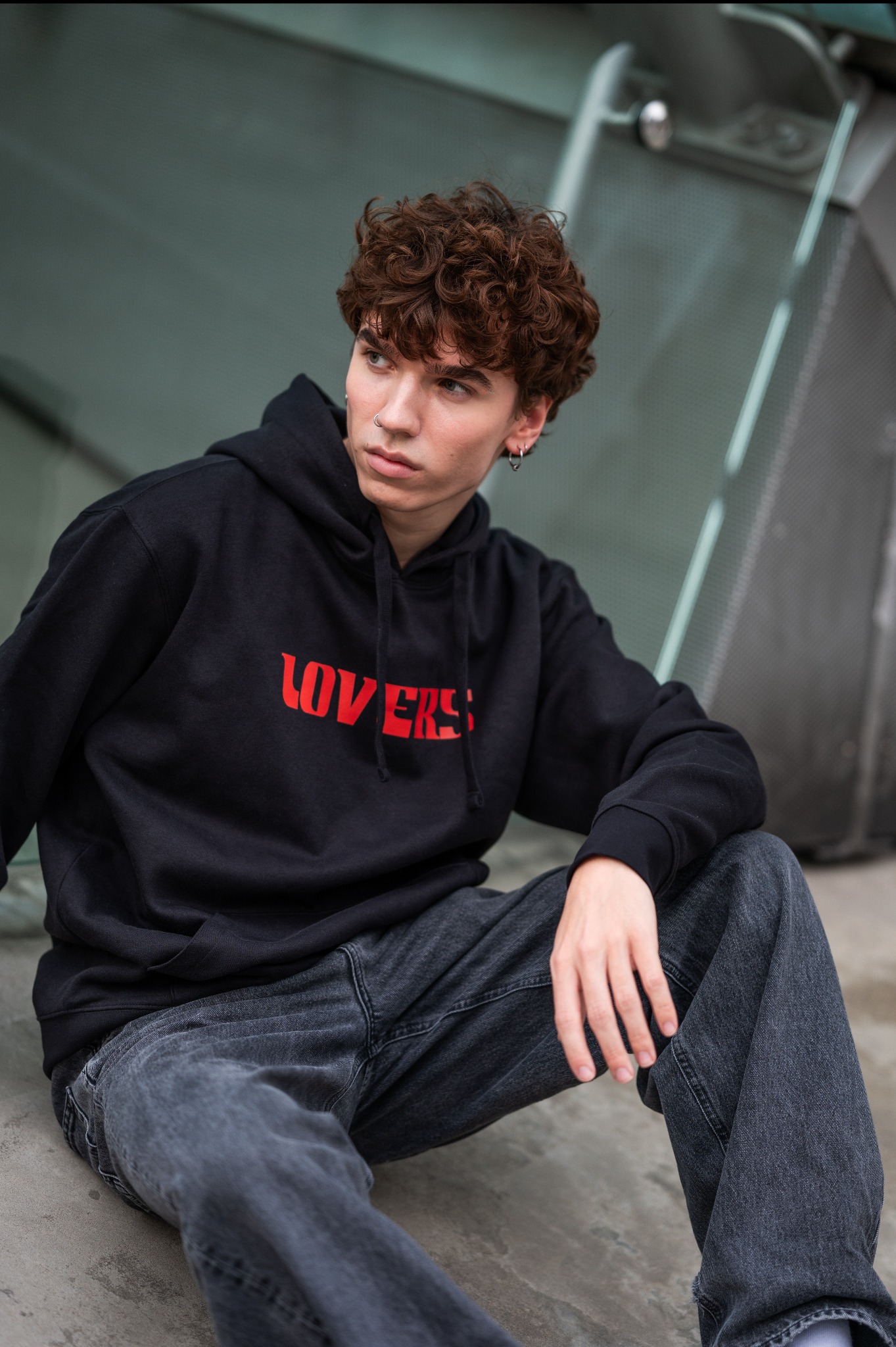 Młody mężczyzna z kręconymi włosami siedzi na tle szklanej ściany, ubrany w czarną bluzę z czerwonym napisem 'LOVERS' i szare dżinsy.