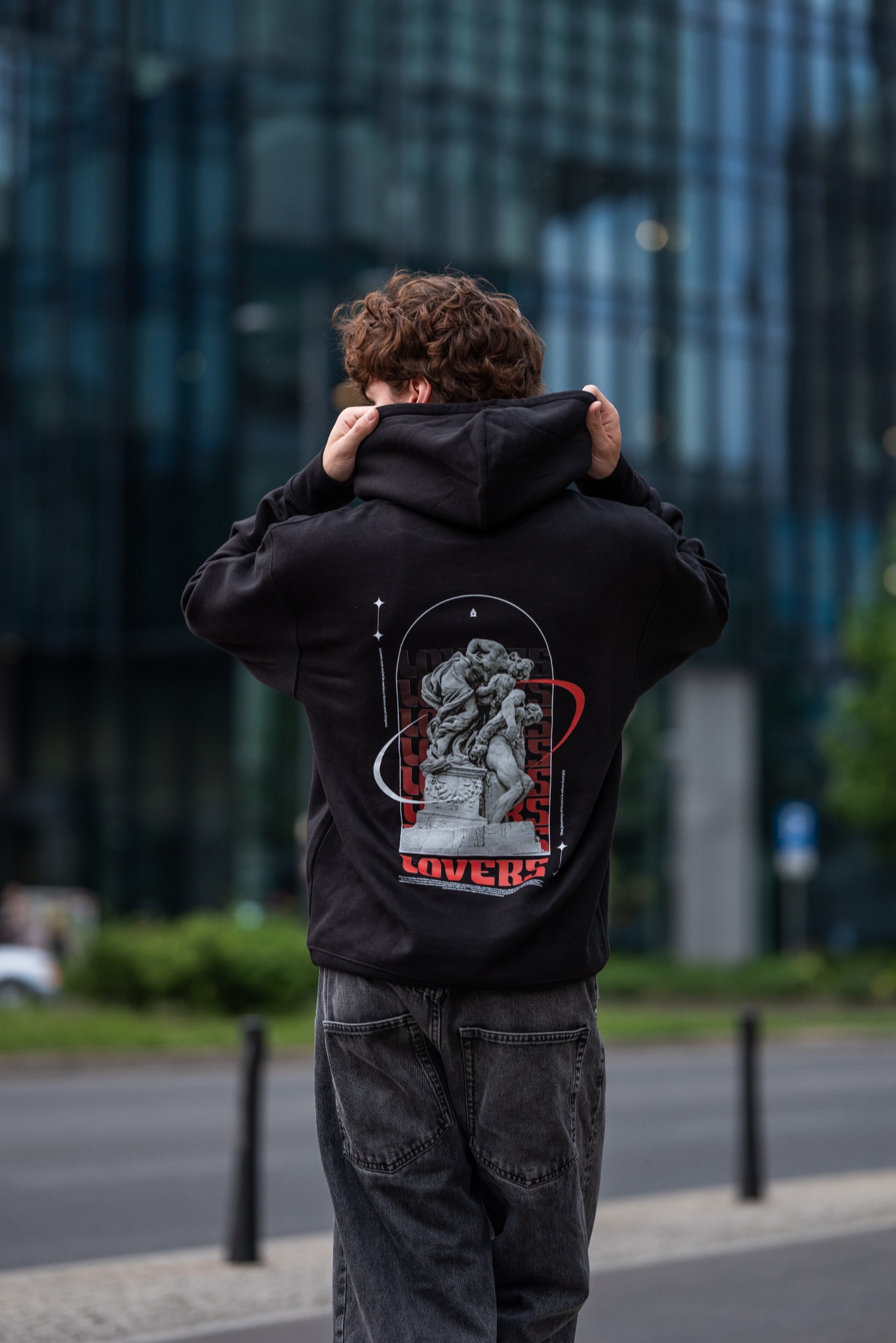 Osoba w czarnej bluzie z kapturem, na której nadrukowana jest statua zakochanych w stylu vintage z napisem Lovers, stojąca tyłem na tle nowoczesnego budynku biurowego.
