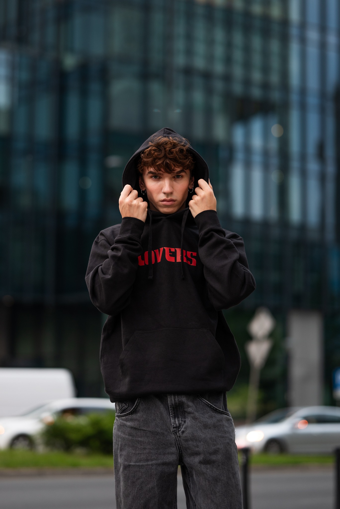 Młody mężczyzna w czarnej bluzie z kapturem i czerwonym napisem 'LOVERS' na tle nowoczesnego budynku biurowego z szklaną fasadą.