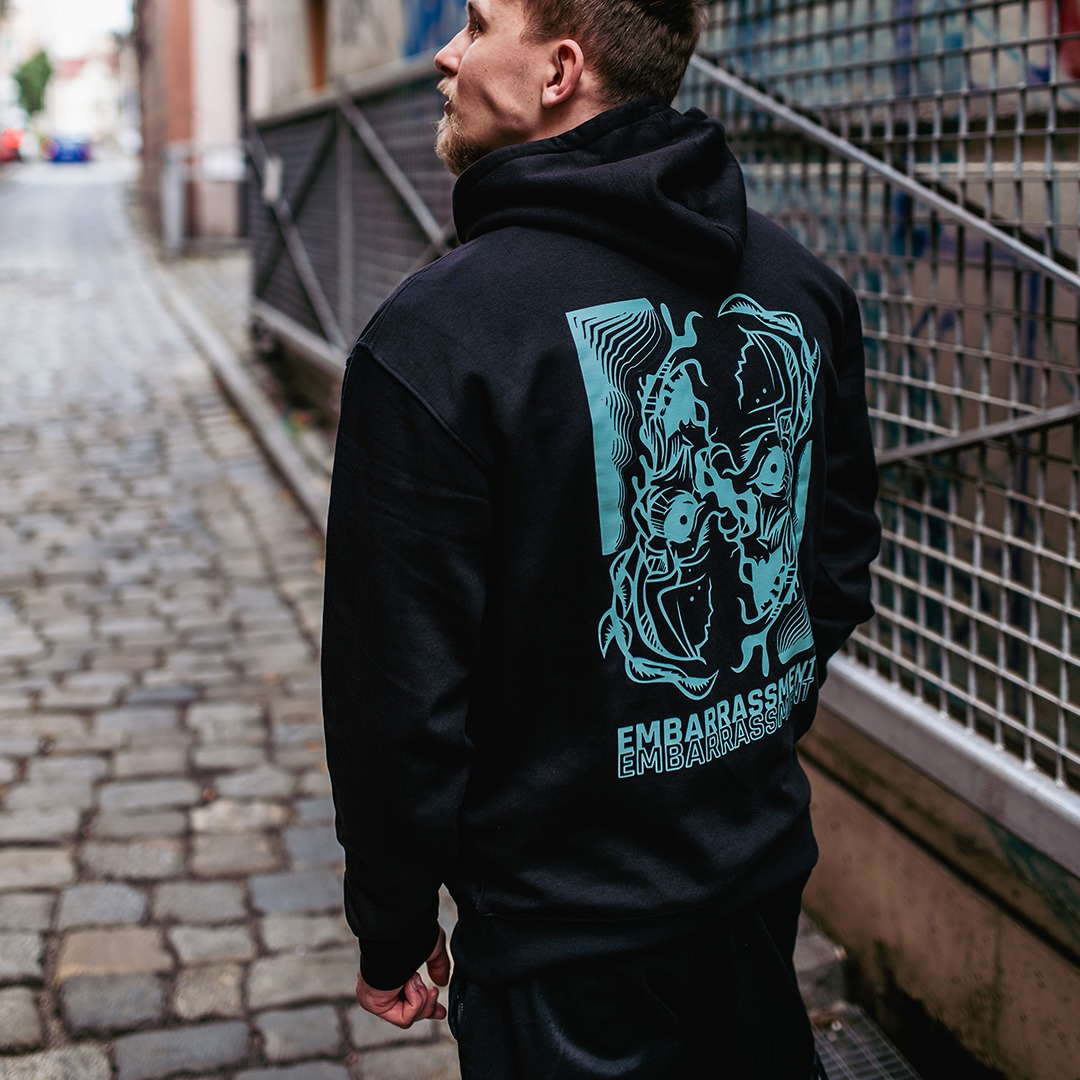 Mężczyzna w czarnej bluzie z kapturem, z turkusowym nadrukiem graficznym na plecach, stojący na brukowanej uliczce w miejskim otoczeniu.