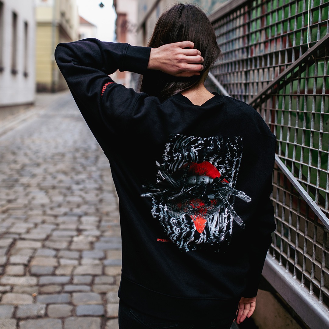 Czarna bluza z nadrukiem graficznym na plecach przedstawiającym abstrakcyjny motyw z czerwonymi akcentami, widoczna na tle uliczki z brukowaną nawierzchnią i metalowej kraty.