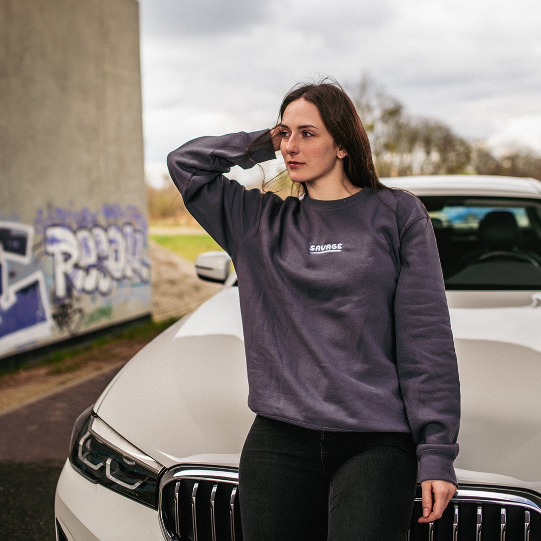 Kobieta oparta o biały samochód w szarej bluzie z napisem 'SAVAGE', w tle betonowy mur z graffiti i pochmurne niebo.