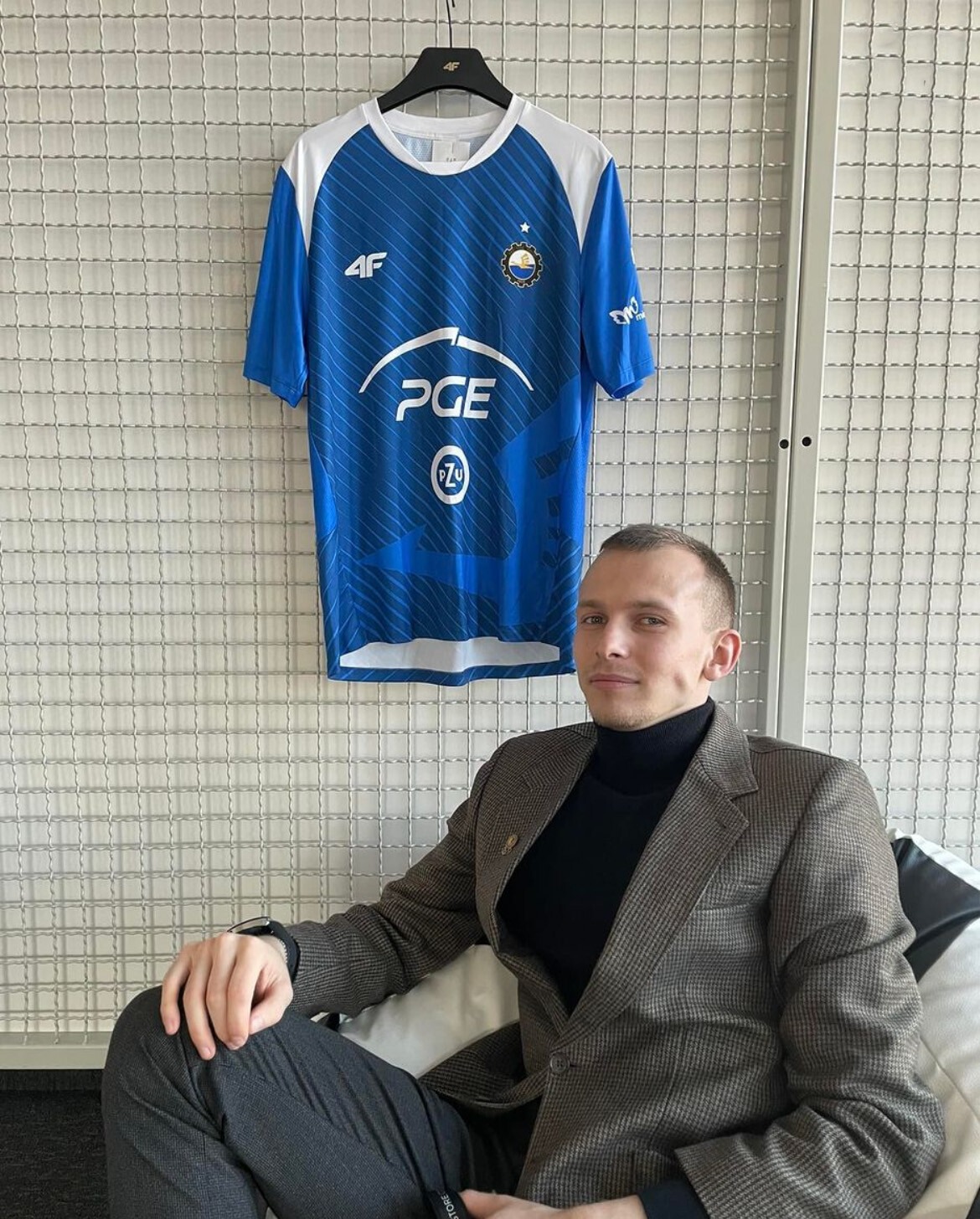 Mężczyzna w tweedowej marynarce siedzi na tle białej ściany z siatki, obok wisi niebieska koszulka sportowa z logo PGE i innymi nadrukami.
