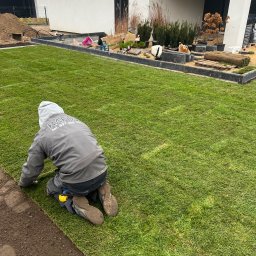 Rafos-garden - Pracownik w szarej bluzie z kapturem i logo firmy układa rolki trawy na przygotowanym podłożu, w tle nowoczesny dom z elementami dekoracyjnymi i roślinnością.