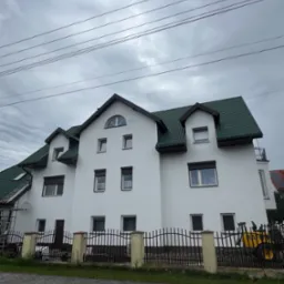 Biały, dwupiętrowy dom z zielonym dachem, widziany z poziomu chodnika. Metalowe ogrodzenie z betonowymi słupkami. Pochmurne niebo w tle. Widoczne przewody elektryczne.