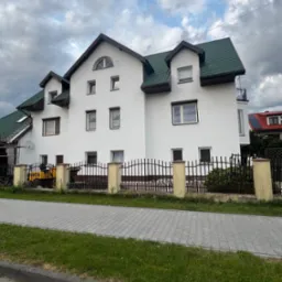 Dwupiętrowy dom z zielonym dachem i białą elewacją, otoczony metalowym ogrodzeniem z betonowymi słupkami. Na podwórku widoczna żółta maszyna budowlana.