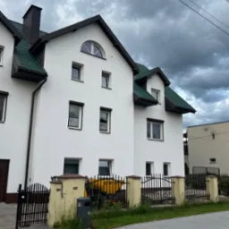 Biały dom z zielonym dachem, widoczny z poziomu chodnika. W tle pochmurne niebo i słupy elektryczne. Żółty sprzęt budowlany za ogrodzeniem.