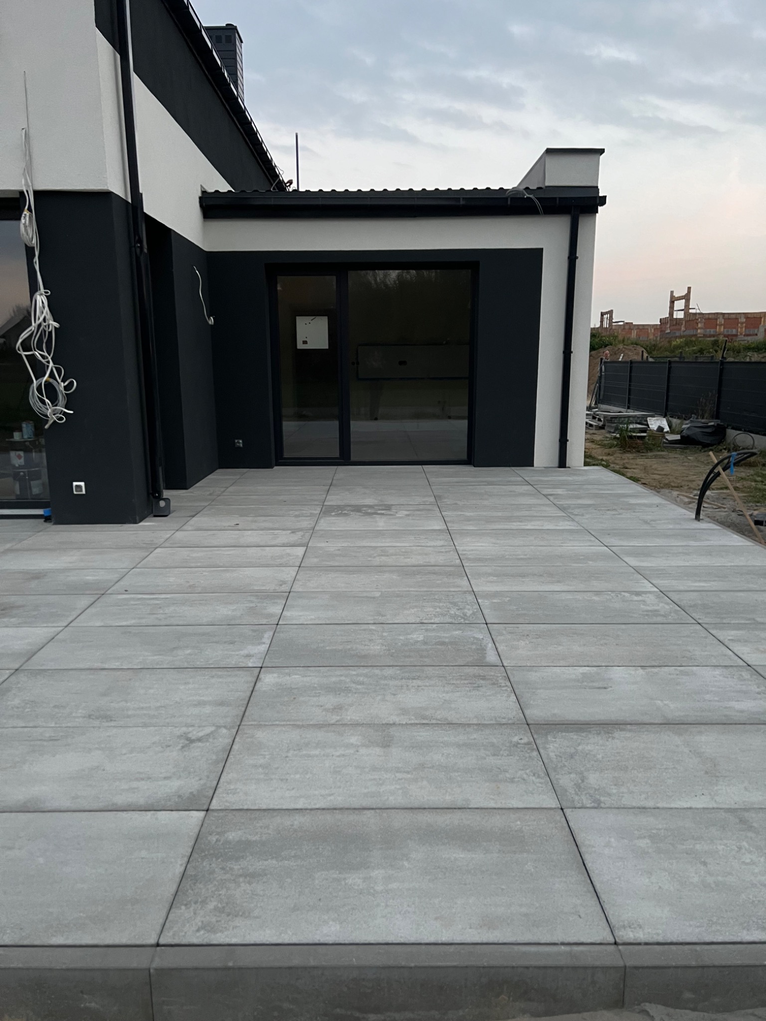 Nowoczesny taras wyłożony szarą kostką brukową w Sieradzu, z widokiem na minimalistyczny budynek z czarno-białą elewacją i przeszkleniami.